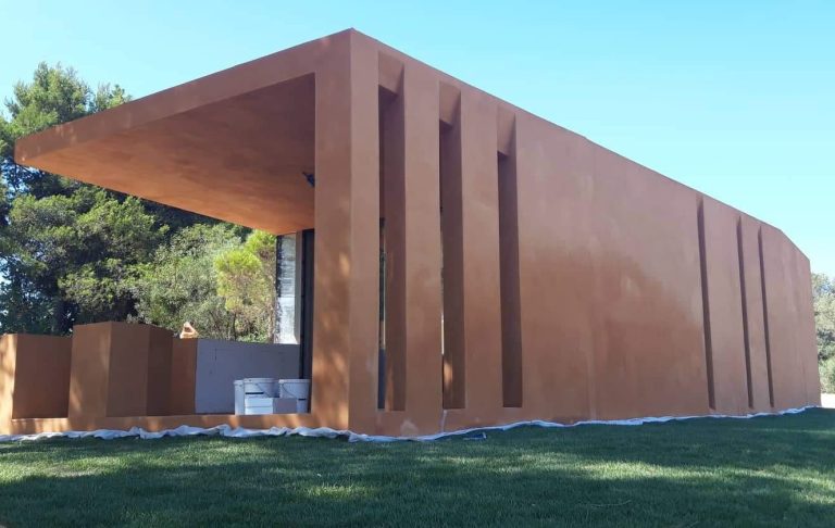 Fotografía de un pabellón moderno de un solo piso con revestimiento texturizado de arcilla terracota. La fachada principal tiene aberturas verticales estrechas y un gran voladizo cubierto a la izquierda. Se encuentra sobre césped con lonas de protección, rodeado de un bosque de pinos y bajo un cielo azul despejado.