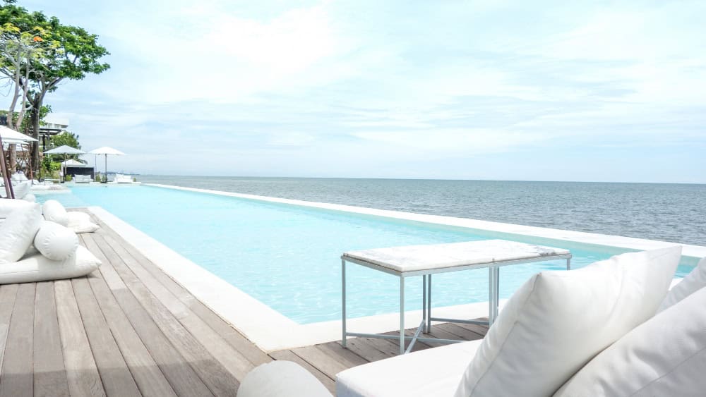Detalle del borde de una piscina exterior con revestimiento natural mineral de Naturclay, junto a una tarima de madera con cojines blancos frente al mar.